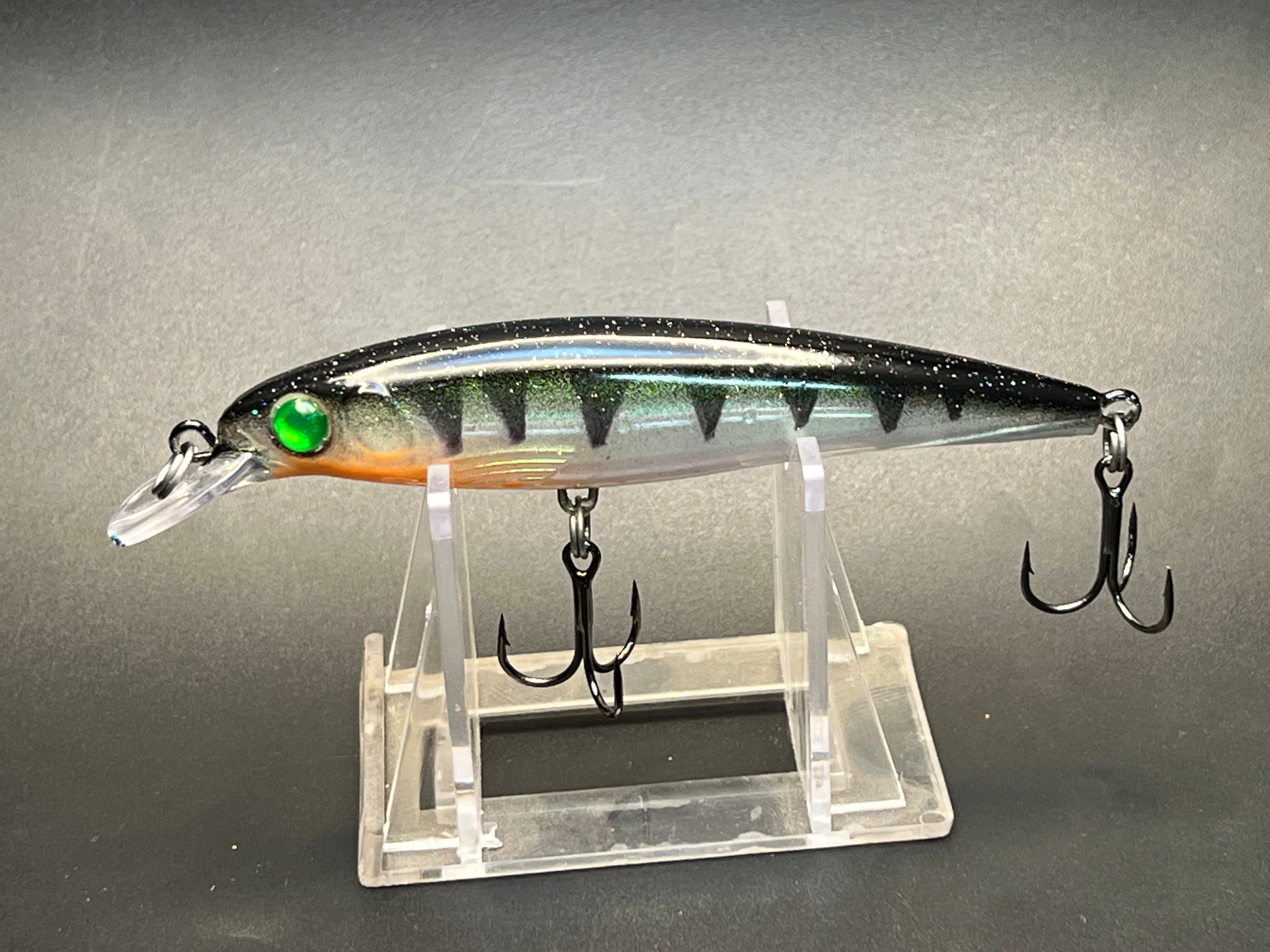 Fishing lure with black body, green eye, and treble hooks on clear stand