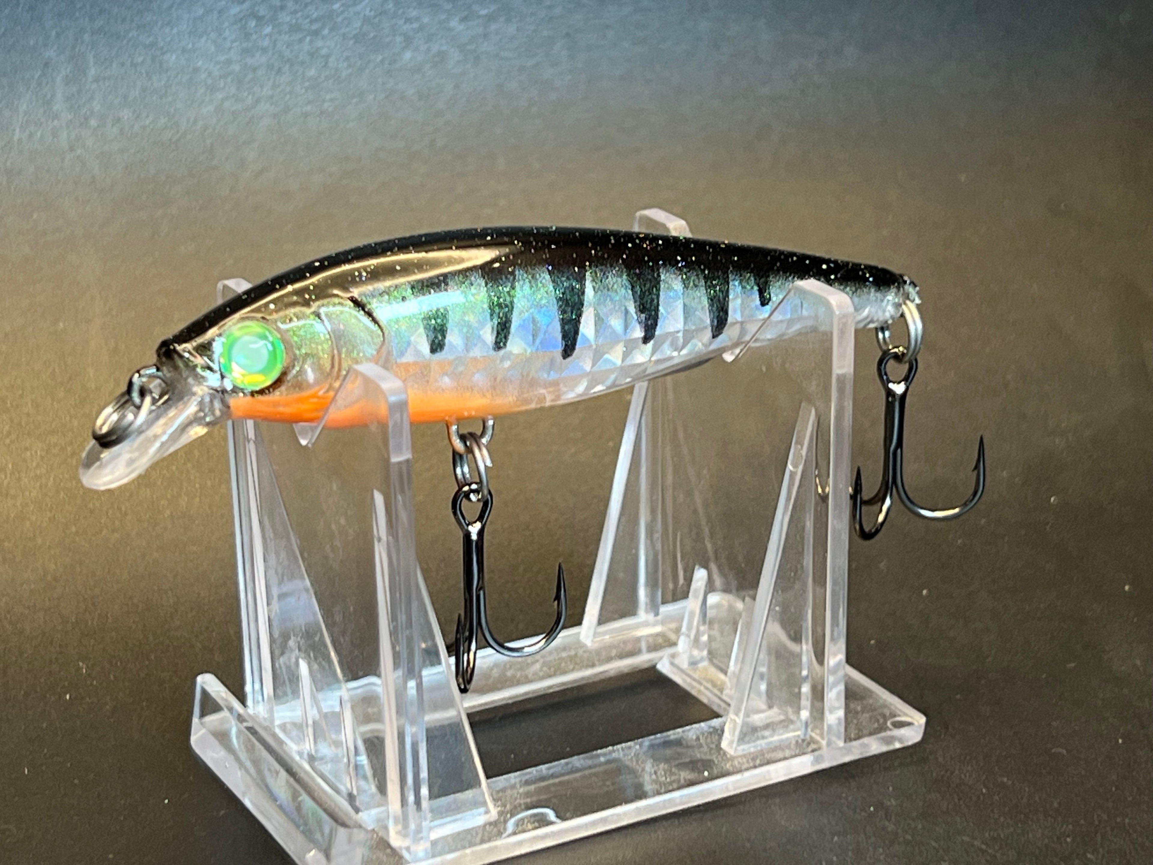 Iridescent silver fishing jerkbait with orange stripe, green eye, and black treble hooks