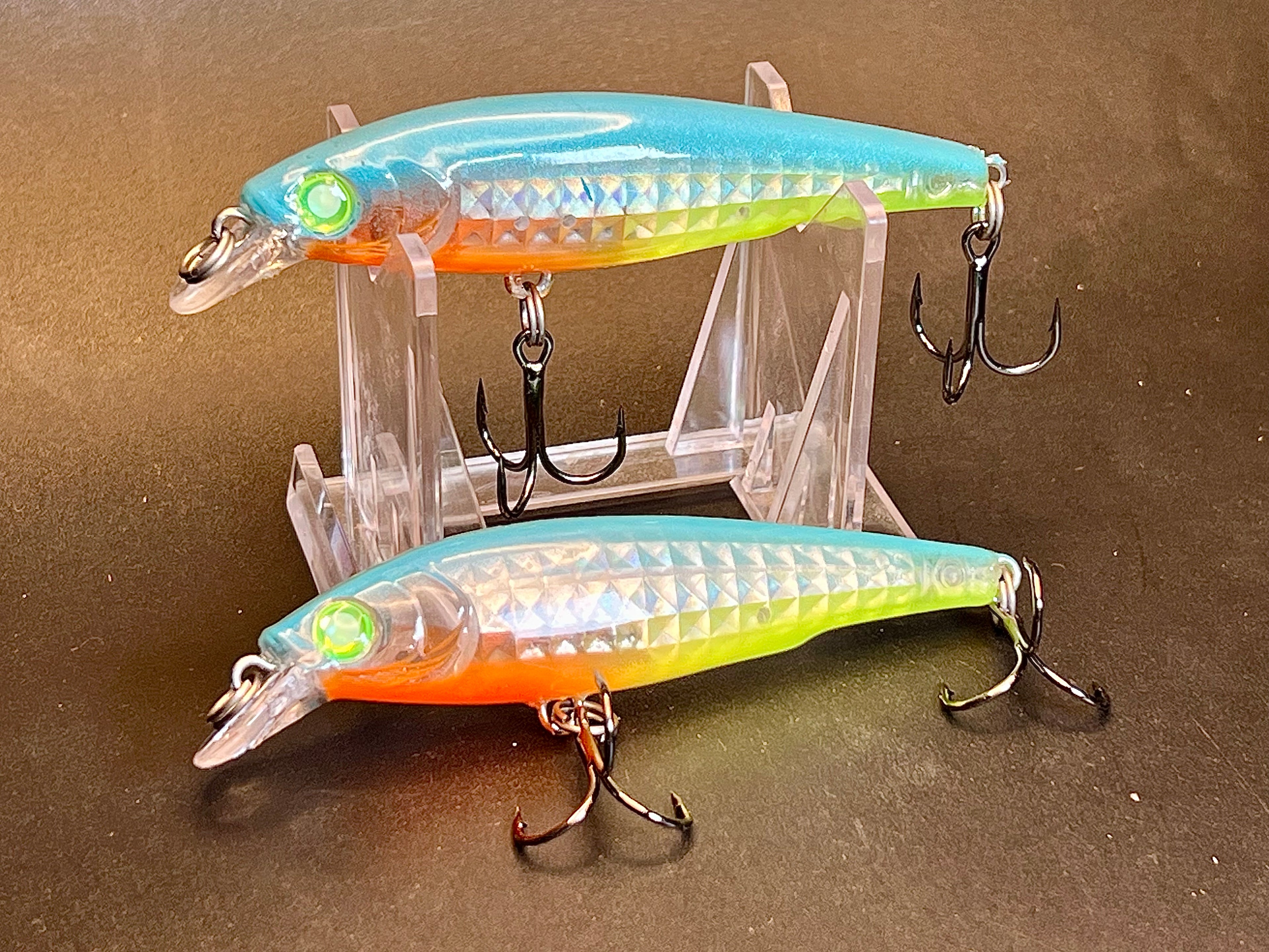 Two metallic orange and chartreuse fishing jerkbaits on clear stand