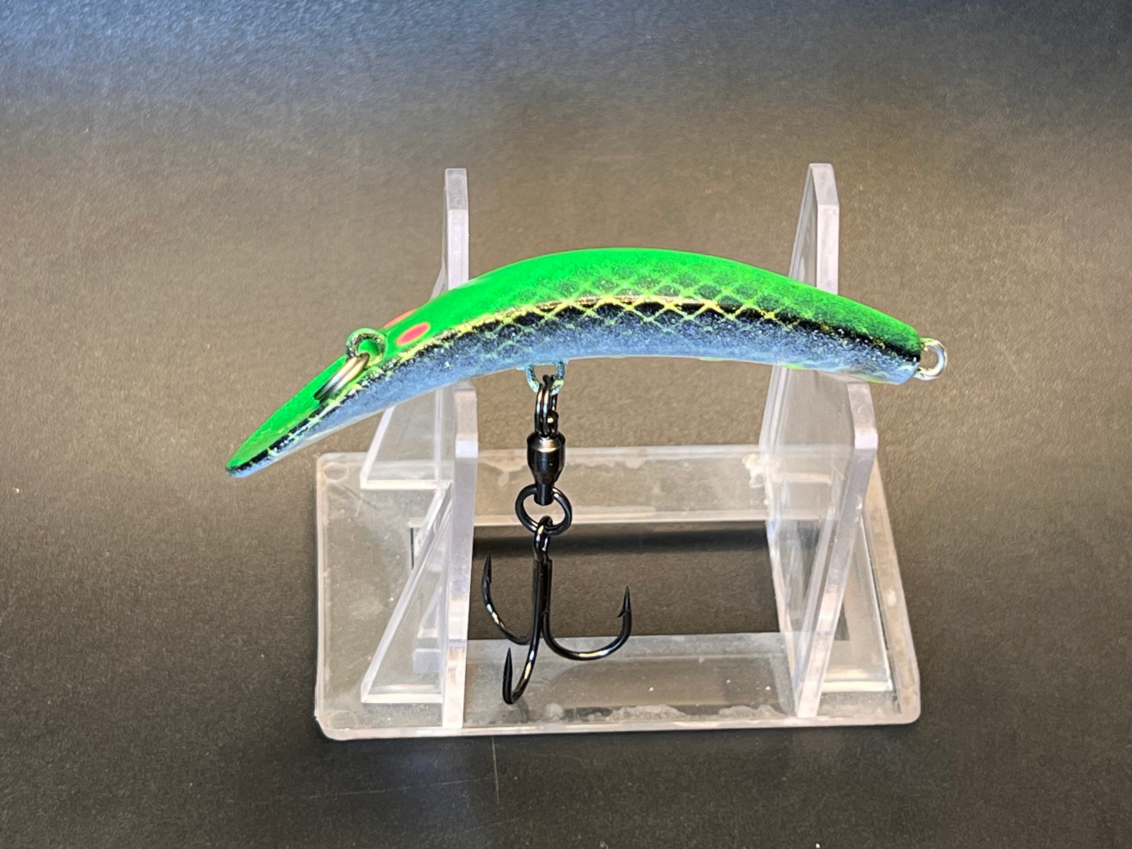 Green fish-shaped fishing lure with blue edge and two treble hooks on display stand