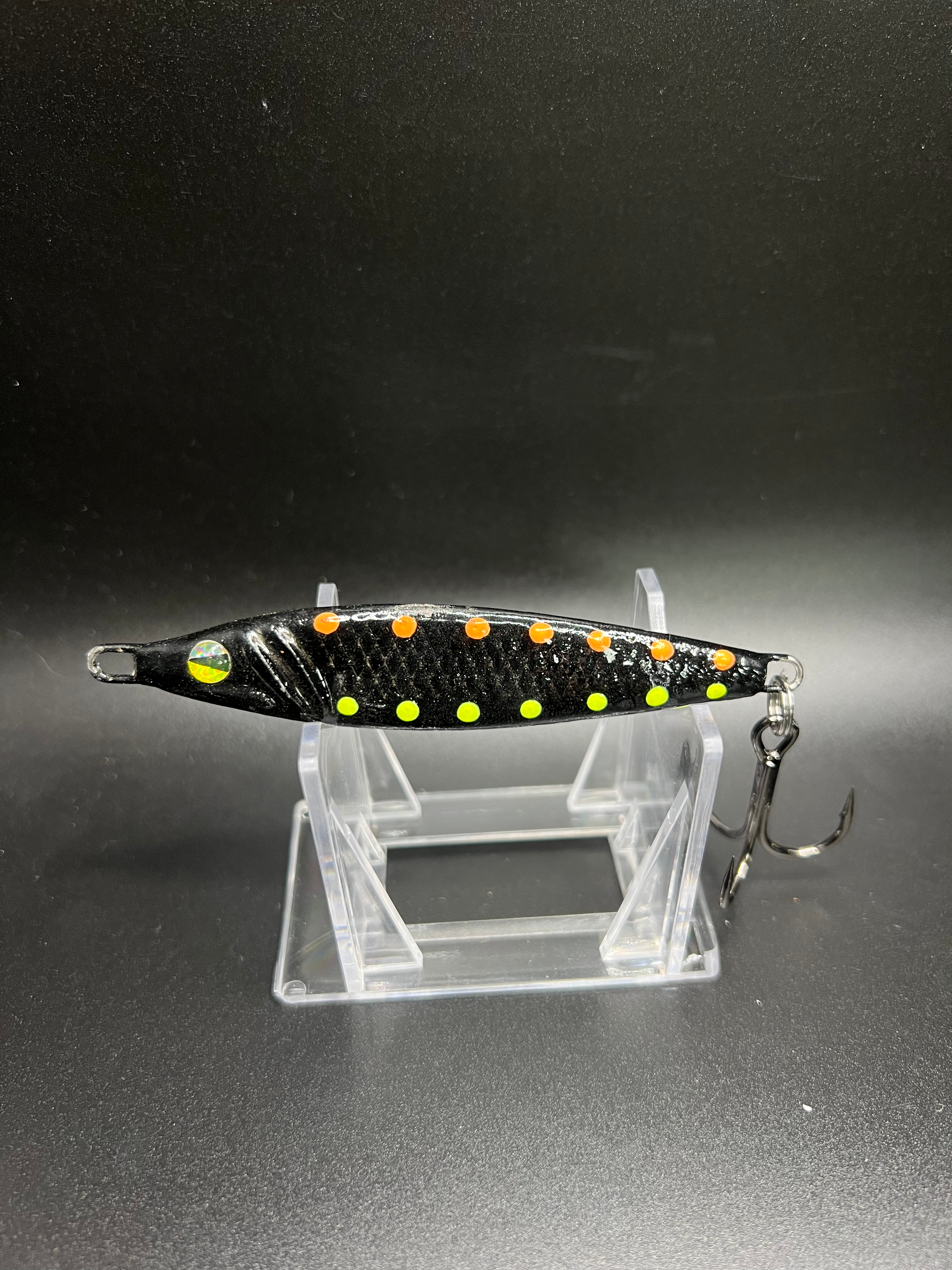 2 oz jigging spoon with black body, orange/yellow dots, holographic eye and treble hook