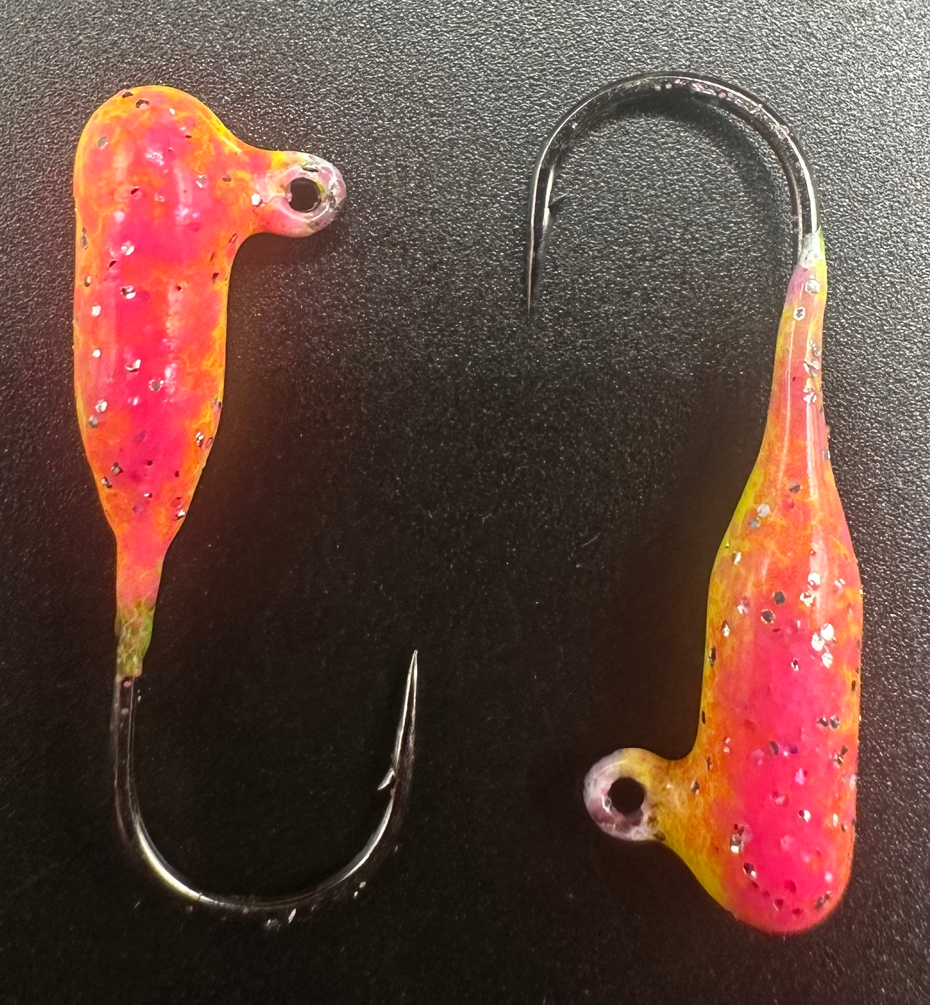 Two tube jigs on dark surface: pink and orange glow, speckled, curved hooks